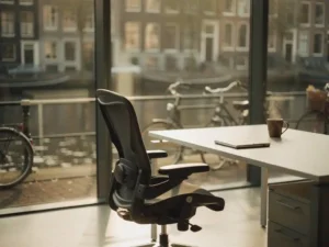 Ergonomische bureaustoel bij een modern werkstation in Amsterdam, dampende koffiemok naast een notitieboek, ochtendlicht door hoge ramen met uitzicht op een gracht.