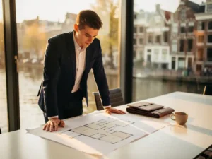 Jonge professional in zakelijk pak bestudeert architectonische plattegrond in vergaderzaal met uitzicht op Amsterdam.